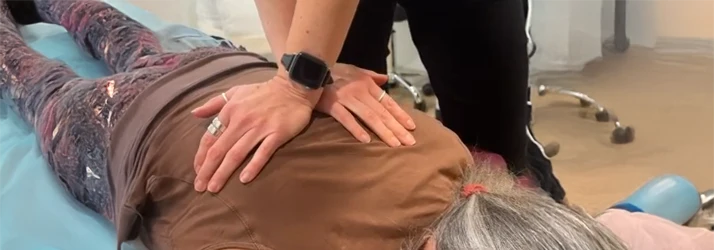 A clinician performs a manual spinal adjustment on an individual lying face‑down on a table.