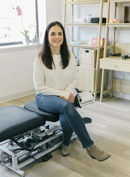 Dr. Ali Perez seated on a chiropractic table in a bright, organized treatment room.