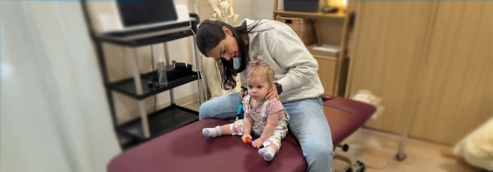 Dr. Ali Perez assists a seated infant on a treatment table during a gentle pediatric care session.